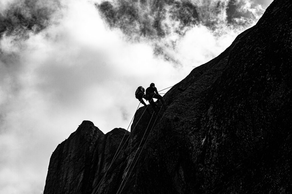 Dramatic black and white photo of climbers ascending a steep cliff against a cloudy sky.