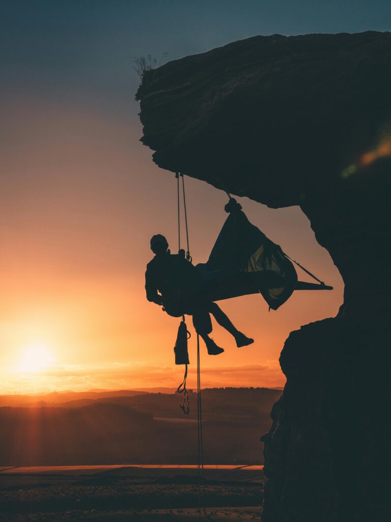 Silhouette of a rock climber hanging from a cliff edge during a colorful sunset, embodying adventure and exploration.